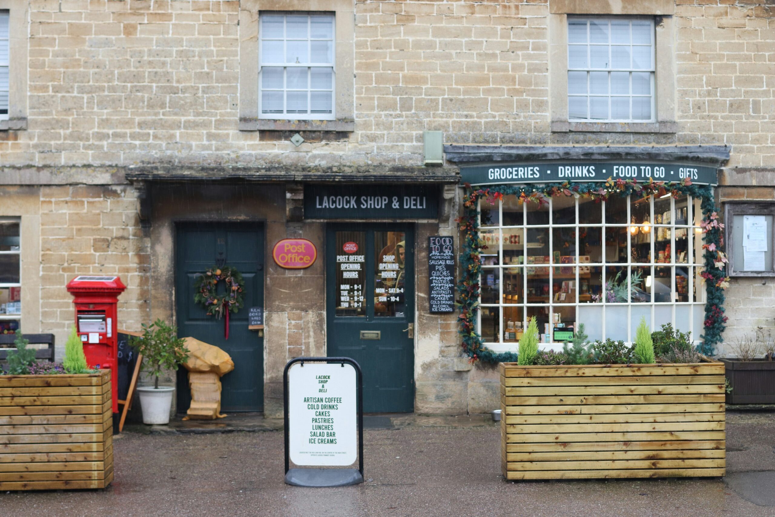 A stone building with a post office and deli; a red postbox, festive wreaths, plants, and a signboard are outside. The deli window displays groceries and drinks.