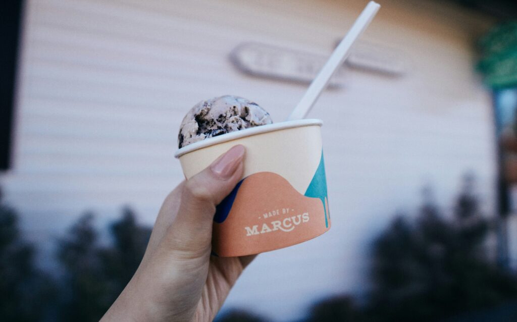 A hand holds a cup of ice cream with a spoon; the cup says Made by: Marcus. The background is blurred with an indistinct building and sign.