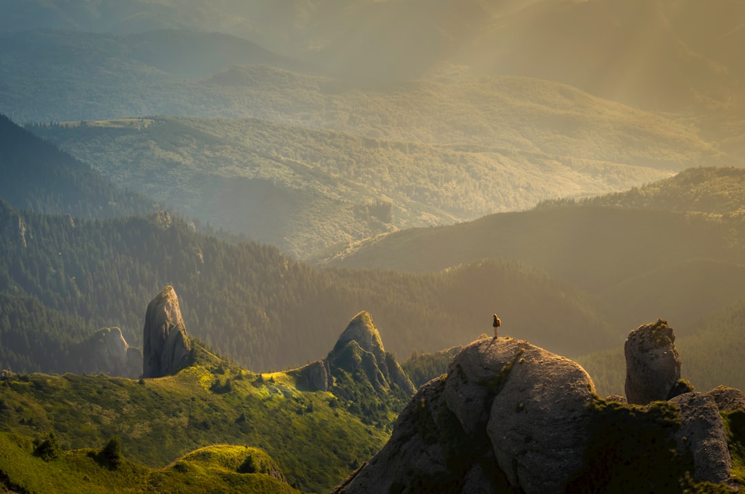 260720261785093919.jpeg A person stands on a rocky mountain peak surrounded by green hills and sunlit valleys, with distant forested mountains fading into the misty background.