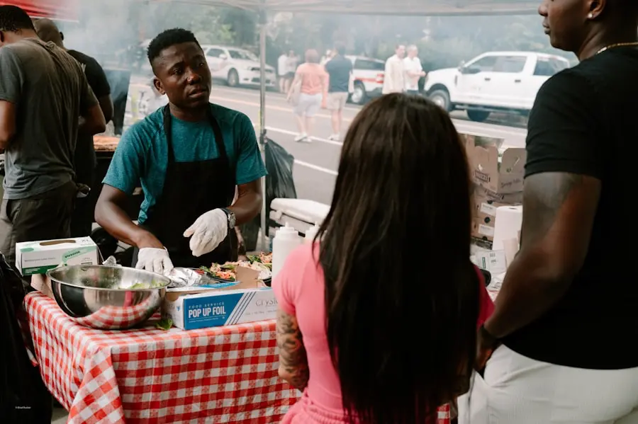 A man wearing gloves and an apron serves food at an outdoor market stall with a red checkered tablecloth, while two people stand across from him. Cars and people are visible in the background. A man wearing gloves and an apron serves food at an outdoor market stall with a red checkered tablecloth, while two people stand across from him. Cars and people are visible in the background.