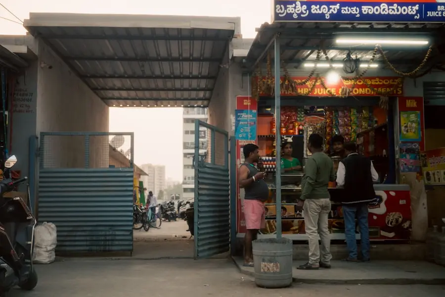A small juice and snack shop with several people gathered at the counter; open gates, bikes, and buildings are visible in the background, and a scooter is parked nearby.