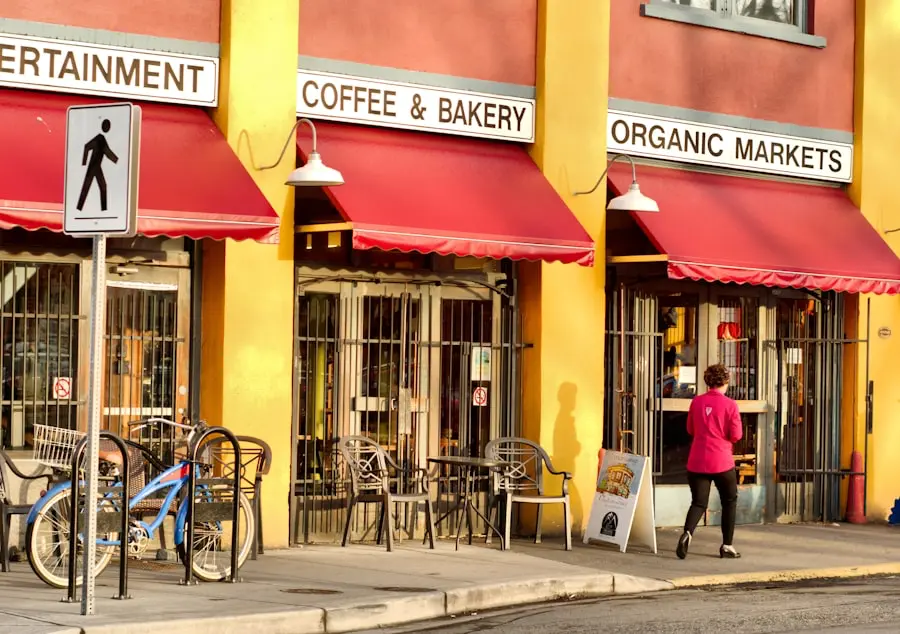 A person walks past a coffee and bakery shop with red awnings and outdoor seating; a bike is parked nearby, and a pedestrian crossing sign is visible in front. A person walks past a coffee and bakery shop with red awnings and outdoor seating; a bike is parked nearby, and a pedestrian crossing sign is visible in front.