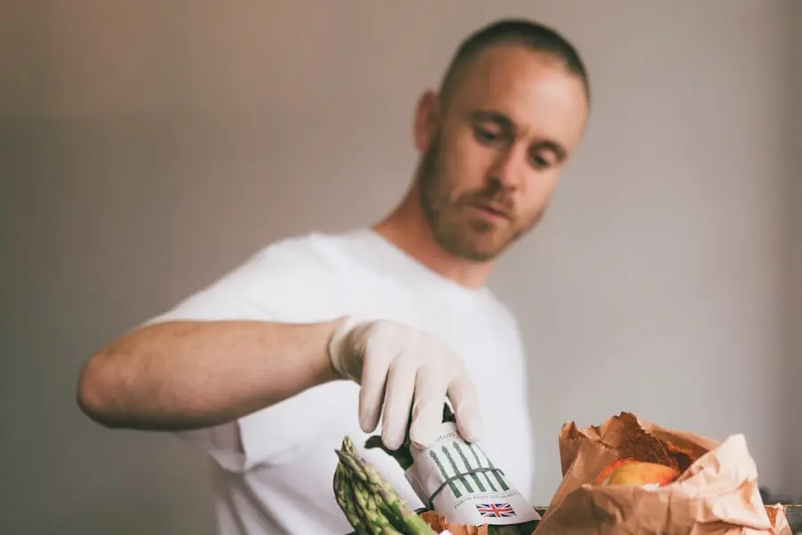 A person wearing a white shirt and gloves holds a can with a UK flag, surrounded by groceries including asparagus and apples in a paper bag, against a neutral background. A person wearing a white shirt and gloves holds a can with a UK flag, surrounded by groceries including asparagus and apples in a paper bag, against a neutral background.