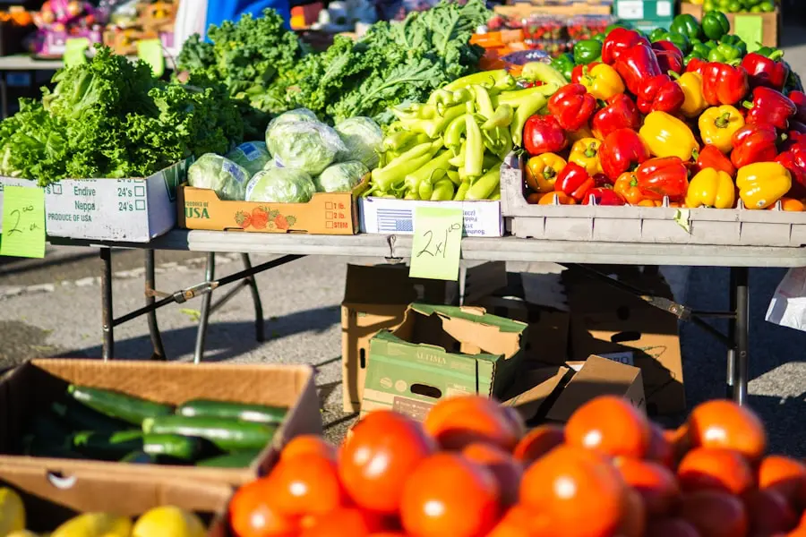 Colorful vegetables including peppers, lettuce, cucumbers, and tomatoes displayed on tables and in boxes at an outdoor farmers market. Handwritten price signs are visible on some produce.