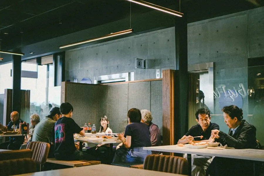 People are sitting at tables in a modern restaurant, eating and talking. The room has concrete walls, large windows, and hanging lights, creating a casual and relaxed atmosphere. People are sitting at tables in a modern restaurant, eating and talking. The room has concrete walls, large windows, and hanging lights, creating a casual and relaxed atmosphere.