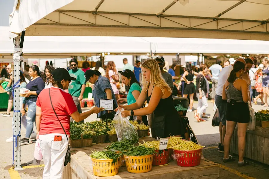 A bustling outdoor farmers market with people shopping for fresh produce, including green beans. A vendor hands a customer a bag of vegetables under a white canopy.