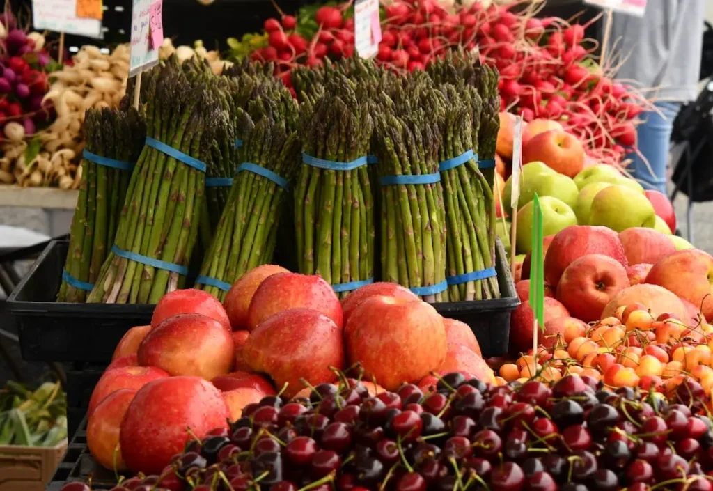 A colorful market display with bundles of asparagus, red apples, green apples, cherries, radishes, and peaches arranged neatly on a table.